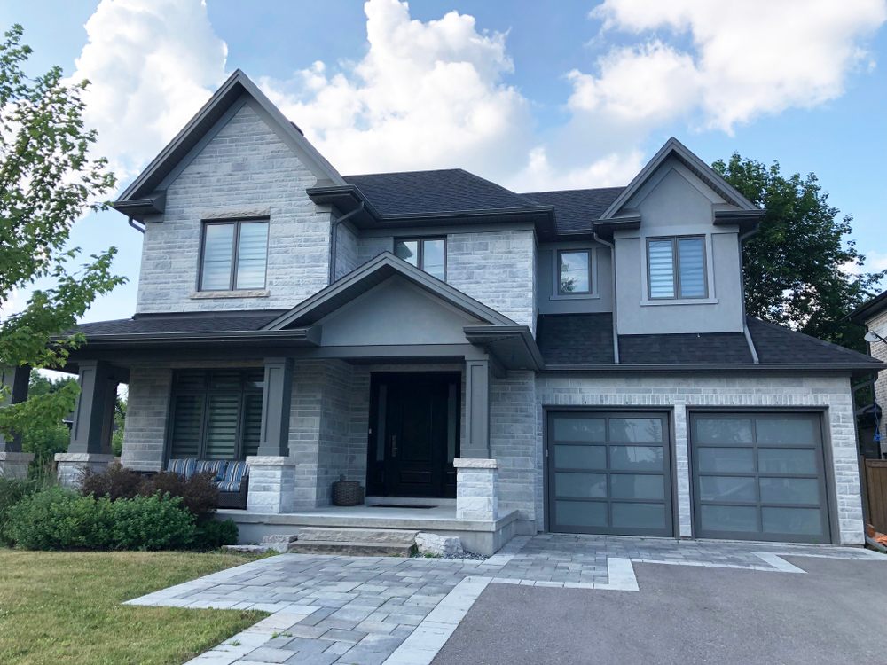 The Ultimate Guide to Interlocking Driveway Designs for Modern Homes. A beautiful, large, modern custom residential house with grey stone and brick, with gray shingles and black and grey trim, patterned interlock walkway and driveway, and double glass garage doors.